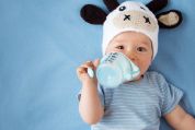 Baby mit gehäkelter Kuhmütze liegt auf Wickeltisch und trinkt einhändig aus Plastikgefäss, von oben betrachtet Baby mit gehäkelter Kuhmütze liegt auf Wickeltisch und trinkt einhändig aus Plastikgefäss, von oben betrachtet